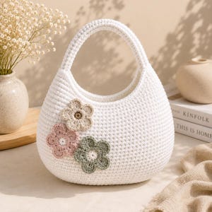 May include: A white crochet handbag with a rounded shape and a single integrated handle. The front of the bag is decorated with three small, flat, crocheted flower appliques in muted shades of beige, dusty pink, and sage green.