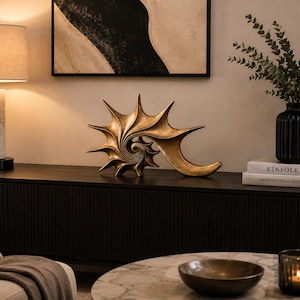 May include: A decorative sculpture shaped like a spiky, spiraling seashell with a metallic gold finish. The object features a complex, curved form with multiple pointed protrusions radiating from a central, tightly coiled spiral center point.