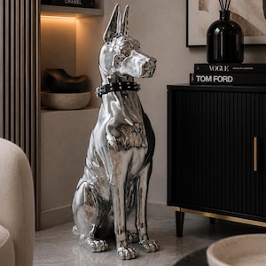 Peut inclure: Une grande sculpture argentée réfléchissante représentant un chien Doberman assis en position verticale. La sculpture présente une finition métallique lisse et est ornée d'un collier noir à clous autour du cou, ajoutant un détail distinct.