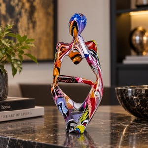 May include: A stylised abstract sculpture of a seated figure with a smooth, glossy finish. The surface features a vibrant, multi-coloured graffiti-style pattern with swirls of blue, red, orange, yellow, black, and white paint across the entire form.