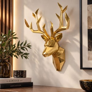Può includere: Una scultura da parete a forma di testa di cervo geometrica color oro, caratterizzata da un design angolare sfaccettato con corna prominenti. La finitura metallica riflette la luce, creando riflessi nitidi e ombre profonde sulla superficie.
