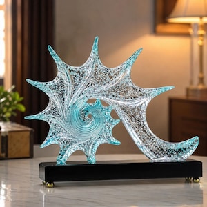 Peut inclure: Une sculpture décorative en verre représentant une forme de coquillage en spirale en forme d'étoile, d'un bleu clair translucide avec des détails de bulles internes. L'objet est monté sur une base noire rectangulaire avec de petits pieds dorés.