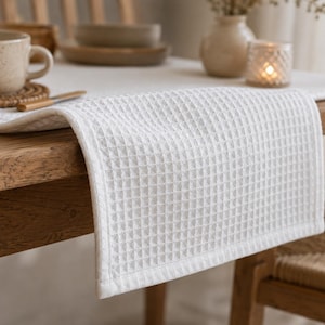 Op de afbeelding: Een witte rechthoekige tafelloper met een gestructureerd wafelpatroon. De stof hangt over de rand van een houten tafel en toont de rasterachtige structuur van het textielmateriaal en de strakke, afgewerkte zomen aan de randen.
