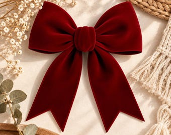 May include: A deep red velvet bow featuring two large upper loops and two long, downward-pointing tails with V-shaped notched ends. The fabric has a smooth, soft texture with a rich, saturated color and a central knot detail connecting the loops.