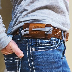 May include: A brown leather knife sheath with a silver folding knife clipped inside, attached to a brown leather belt. The belt is worn with blue jeans. The sheath has a decorative design.