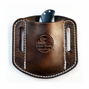 May include: A brown leather knife sheath with white stitching and a black knife partially inserted. The sheath features a circular embossed logo reading "Evil Duck Studios". Two vertical slots are visible on the sides.
