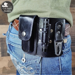 Puede incluir: Funda de cinturón de cuero negro con linterna y bolsillo. La funda está sujeta a un cinturón de cuero marrón y se asegura con remaches plateados. Un mosquetón plateado está sujeto a la funda.