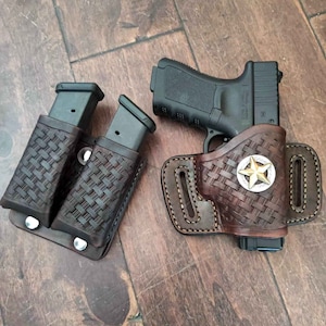 Könnte beinhalten: Ein braunes Lederholster mit Sternabzeichen und passendem Magazinhalter. Das Holster und der Magazinhalter haben ein gewebtes Muster. Eine schwarze Schusswaffe befindet sich im Holster.