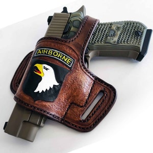 May include: A brown leather holster with the word "AIRBORNE" and a white eagle patch. The holster holds a green and black handgun. The leather has a textured appearance and is stitched along the edges.