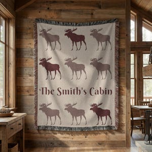 May include: A woven throw blanket featuring a repeating pattern of nine dark brown moose silhouettes arranged in three rows on a light beige background, with the text "The Smith's Cabin" printed in a dark brown script font across the centre.