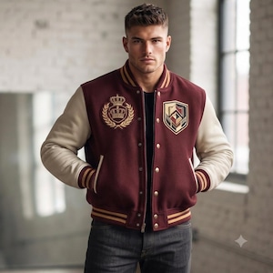 May include: A burgundy varsity jacket with cream-colored sleeves, featuring a gold embroidered crown emblem on the right chest and a geometric shield patch on the left. The jacket has a snap-button front, ribbed striped cuffs, and a ribbed hem.