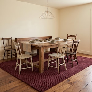 Puede incluir: Una mesa de comedor de madera con un banco largo y cinco sillas, incluyendo dos sillas claras y tres oscuras. La mesa cuenta con un camino de mesa beige y varios cuencos, situada sobre una alfombra grande de color rojo oscuro estriado.