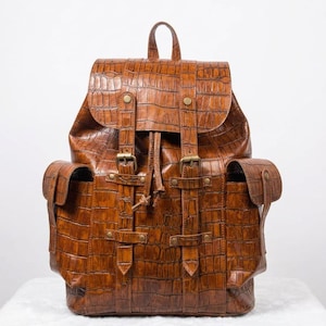 May include: A brown backpack with a textured crocodile-embossed pattern. The bag features a top flap with two vertical straps, brass-toned metal buckles, and snap closures. It includes two side pockets with flaps and a drawstring main compartment.