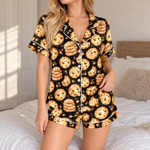 May include: A black pajama set featuring a repeating pattern of golden-brown chocolate chip cookies. The short-sleeved button-down top has a notched collar and white piping, while the matching shorts also feature white piping along the hem.
