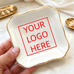 Puede incluir: Un plato pequeño cuadrado de cerámica blanca con bordes ondulados y un borde pintado en oro. El centro del plato presenta un contorno cuadrado rojo que contiene el texto rojo YOUR LOGO HERE. El plato tiene un acabado liso y brillante.