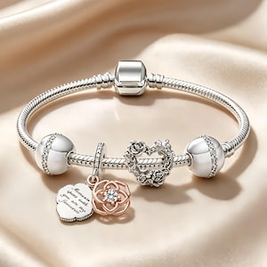 Könnte beinhalten: Ein silbernes Schlangenketten-Armband mit zwei silbernen Kugeln mit klaren Steinen, einem herzförmigen Anhänger mit Rosen-Details und zwei hängenden Charms: einer roségoldenen Blume mit einem Stein und einem silbernen Anhänger mit Text.
