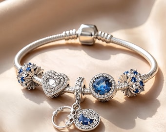 Op de afbeelding: Een zilveren bedelarmband met een centraal hartvormig bedeltje met heldere stenen, een ronde bedel met een diepblauwe steen, twee bolvormige bedels met blauwe en heldere stenen, en een hangende bedel met een halve maan en sterren.