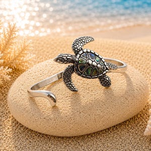 Könnte beinhalten: Ein silberfarbenes Armband mit einer Meeresschildkröte als Mittelstück. Der Panzer der Schildkröte ist mit schillernden Abalone-Muschelsegmenten in Grün- und Blautönen eingelegt. Der Metallkörper der Schildkröte hat eine texturierte Oberfläche.