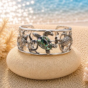 May include: A silver-toned cuff bracelet featuring an openwork design of multiple sea turtles. The central turtle is inlaid with a polished, iridescent abalone shell piece displaying shades of green, blue, and purple against the metallic frame.