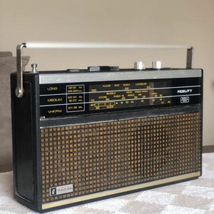 May include: A black and silver Fidelity RAD 26 portable radio featuring a brown grid-patterned speaker grille, a top-mounted carrying handle, and a black control panel with white text displaying frequency bands for Long, Medium, and VHF/FM.
