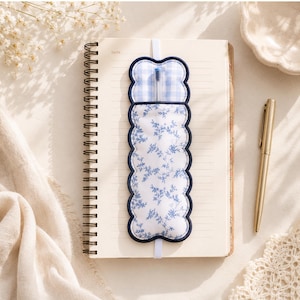 Puede incluir: Un portalápices de tela con bordes festoneados, que presenta un estampado floral azul y blanco en la sección inferior y un patrón de cuadros vichy azul y blanco en la superior, sujeto a un cuaderno de espiral con una banda elástica blanca.