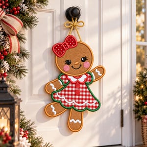 May include: A gingerbread person door hanger featuring a brown cookie-shaped body with white icing details, wearing a red and white gingham dress with a green trim, and a red bow with white polka dots on its head, suspended by a gold cord.