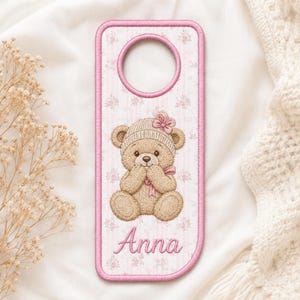 May include: A rectangular door hanger with rounded corners and a circular cutout at the top. The fabric features a light pink striped and floral pattern with a central embroidered tan teddy bear wearing a pink bow and the name Anna in pink.