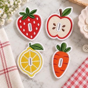 May include: Four embroidered patches shaped like a strawberry, a sliced apple, a lemon slice, and a carrot. Each patch features a central vertical white oval cutout. The strawberry is red with white seeds, the apple is cream and red, the lemon is yellow, and the carrot is orange with green tops.