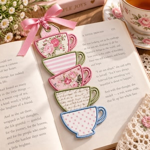 May include: A fabric bookmark featuring a vertical stack of five embroidered teacup shapes in various patterns, including pink roses, white and pink stripes, cursive script, and pink polka dots, topped with a pink satin ribbon bow.
