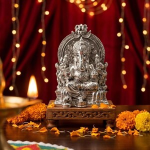 May include: A silver-colored statue of the deity Ganesha seated on an ornate throne, placed upon a rectangular wooden base. The figure features intricate detailing, including a trunk, multiple arms, and a crown, with the text IndianCultureHub visible.