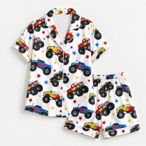 May include: A white short-sleeve button-down pajama shirt and matching shorts set featuring an all-over print of colorful monster trucks in red, yellow, and blue with large black tires, accented by scattered small red, blue, and yellow stars.