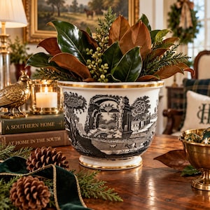 May include: A white ceramic planter featuring a black and white toile-style landscape print of classical ruins and a gold-trimmed rim and base. The planter is filled with a lush arrangement of green magnolia leaves and small green berries.