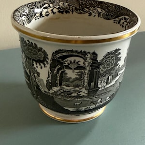 May include: A ceramic planter featuring a black and white transferware pattern of classical ruins, trees, and small figures. The rim and base are accented with a thin gold band, and the interior upper edge displays a decorative floral border.
