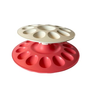 May include: A two-tiered circular egg storage rack featuring a large red base with twelve oval indentations and a smaller cream-coloured top tier with eight oval indentations. The structure is connected by a central pedestal, creating a tiered display.