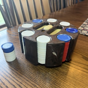 May include: A dark brown marbled plastic carousel poker chip holder containing stacks of white, red, and blue poker chips. A separate small stack of white chips topped with a blue chip sits to the left of the circular rotating storage unit.
