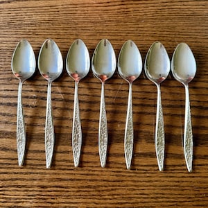 Puede incluir: Un conjunto de siete cucharas de metal plateado dispuestas en fila. Cada cuchara presenta un cuenco ovalado liso y un mango decorado con un patrón geométrico texturizado que consiste en pequeñas líneas angulares y entrecruzadas.
