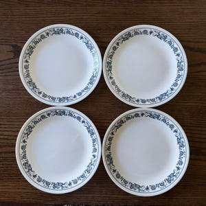 Può includere: Un set di quattro piatti rotondi bianchi, ciascuno caratterizzato da un motivo floreale e a viticci blu scuro continuo lungo il bordo esterno. I piatti presentano un design circolare liscio con un centro bianco e un bordo decorativo.