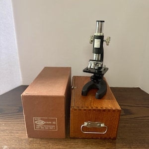 Könnte beinhalten: Ein schwarzes Research Mark XII Revolvermikroskop aus Metall mit silbernem Okular und Objektivlinsen, platziert auf einer hölzernen Aufbewahrungsbox neben einem braunen Karton mit Produktnamen und Vergrößerungsstufen 100-300-400-500 X.