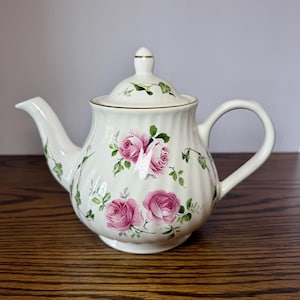 Puede incluir: Una tetera de porcelana color crema con cuerpo acanalado, tapa a juego con pomo y un delicado estampado floral de rosas rosadas con enredaderas de hiedra verde. El borde de la tapa y el pico están decorados con un fino borde dorado.