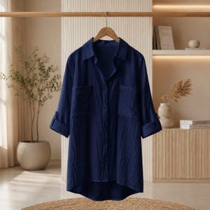 Puede incluir: Una camisa azul oscuro con botones y un acabado de tela texturizada y arrugada. La camisa presenta mangas largas con puños enrollados, un cuello clásico y dos grandes bolsillos de parche en la zona del pecho. Tiene un dobladillo asimétrico.