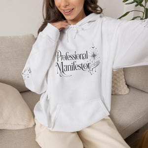 May include: A white hooded sweatshirt featuring a black graphic print on the chest. The text reads Professional Manifestor in a stylized font, accompanied by an illustration of a hand reaching toward a starburst, moon, and celestial elements.
