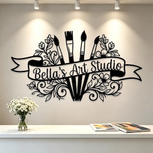 Könnte beinhalten: Ein schwarzes Wand-Schild mit einem zentralen Banner mit dem Text Bella's Art Studio, umgeben von vier Pinseln und komplizierten floralen Schnörkeln. Das Design ist symmetrisch und zeigt künstlerische Werkzeuge sowie botanische Elemente.