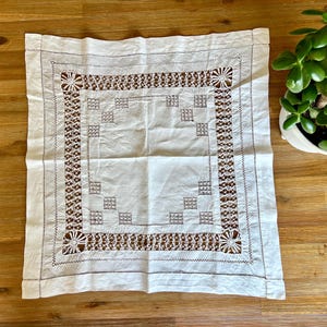 May include: A square white fabric cloth featuring an intricate openwork border with geometric cutouts and circular corner motifs. The central area is plain white fabric, framed by a decorative lattice pattern and small square embroidered accents.