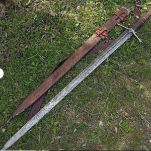 Könnte beinhalten: Ein langes Stahlschwert mit einem sichtbaren wellenförmigen Damastmuster auf der Klinge, ausgestattet mit einem kreuzförmigen Metallgefäß und einem dunkelbraunen, lederumwickelten Griff. Neben dem Schwert liegt eine passende braune Lederscheide.