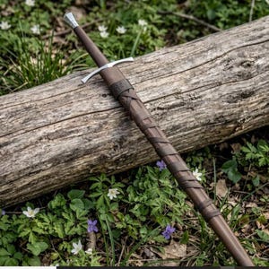 May include: A longsword with a brown leather-wrapped scabbard and hilt. The sword features a silver-coloured metal pommel and a straight crossguard. The scabbard is decorated with a dark brown leather strap wrapped in a crisscross pattern.