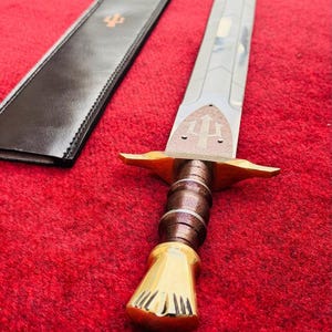 May include: A silver-toned sword with a metallic blade featuring geometric engravings, a brown textured grip, a gold-coloured pommel and crossguard, and a brown leather scabbard with a gold trident emblem embossed on the surface.