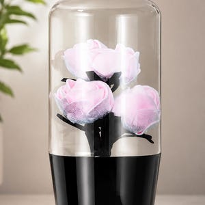 May include: A cylindrical glass cloche with a black base containing three light pink fabric roses on black stems. The clear glass dome covers the flowers, which feature delicate, textured petals. The base is a solid, glossy black material.