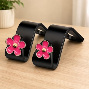 May include: Two black car seat headrest hooks, each featuring a bright pink five-petal flower ornament with a gold-toned center. These S-shaped hooks are designed to attach to the metal posts of a vehicle headrest for hanging bags or items.