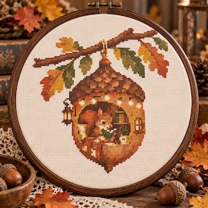 May include: A cross-stitch embroidery in a round wooden hoop featuring an acorn-shaped house hanging from an oak branch. Inside the acorn, a squirrel reads a book. The design includes autumn-colored leaves, a small lantern, and string lights.