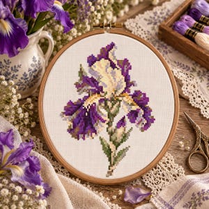 Könnte beinhalten: Ein runder Stickrahmen aus Holz mit weißem Stoff, der ein Kreuzstichmotiv einer einzelnen Irisblüte zeigt. Die Blüte ist in Schattierungen von tiefem Violett, Lila und blassem Gelb gehalten, mit grünen Stängeln und Blättern in der Mitte.